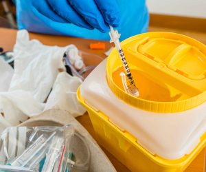 Hand with blue latex glove throws syringe into yellow waste cont
